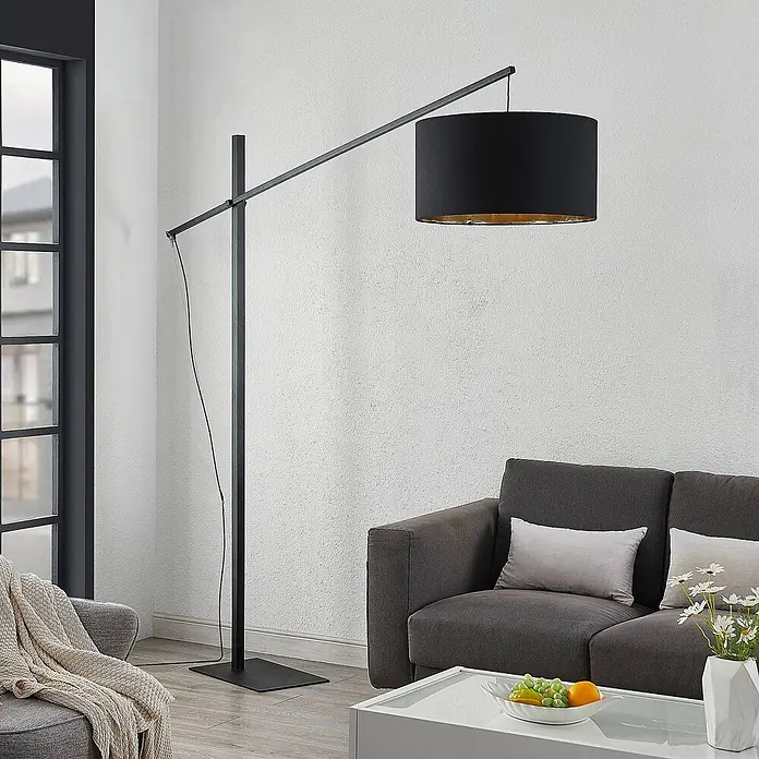 Arcchio Stehleuchte Mossa - schwarz, schwarz, E27 - Moderne schwarze Bogen-Stehleuchte mit zylindrischem Schirm in Schwarz-Gold, über einem dunkelgrauen Sofa im Wohnzimmer.
