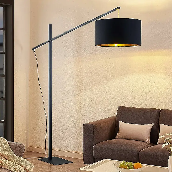 Arcchio Stehleuchte Mossa - schwarz, schwarz, E27 - Schwarze Bogen-Stehlampe aus Metall mit schwarzem Trommelschirm und goldenem Inneren, beleuchtet ein Wohnzimmer neben einem braunen Sofa.