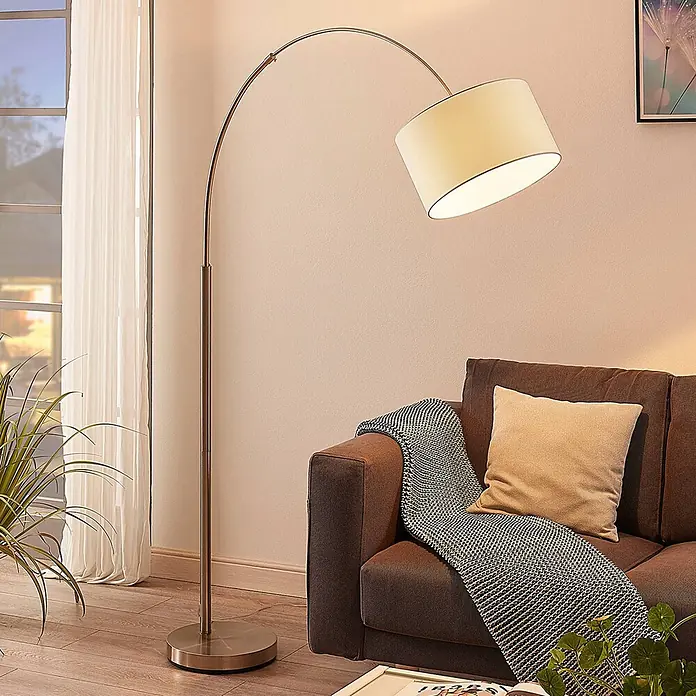 Lindby Stehlampe in nickel satiniert, weiß, Bogenlampe Wohnzimmer -  - Bogenstehlampe mit gebürstetem Metallsockel und cremefarbenem Stoffschirm, die ein Sofa im Wohnzimmer beleuchtet.