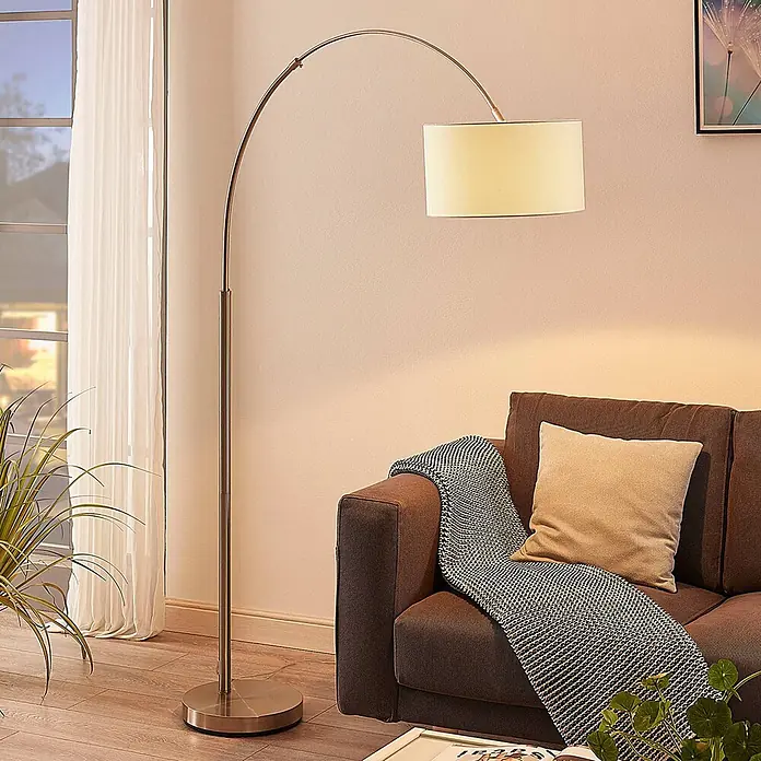 Lindby Stehlampe in nickel satiniert, weiß, Bogenlampe Wohnzimmer -  - Bogenförmige Stehlampe mit gebürsteter Metalloberfläche und beigefarbenem Schirm, spendet warmes Licht neben einem braunen Sofa.