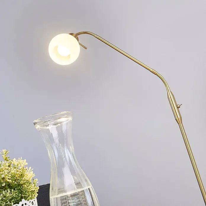 Lindby Stehlampe verstellbarer Glas Leuchtkopf, Standleuchte messing -  - Goldfarbene, flexible Stehleuchte mit kugelförmigem weißem Licht, neben einer Glaskaraffe und Pflanze.