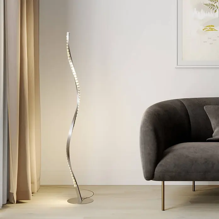 Lindby LED Stehlampe RGB Farbwechsel, dimmbar mit Fernbedienung -  - Eine moderne, geschwungene silberne LED-Stehleuchte steht auf hellem Holzboden und beleuchtet den Bereich neben einem dunkelgrauen Sofa und hellen Vorhängen.