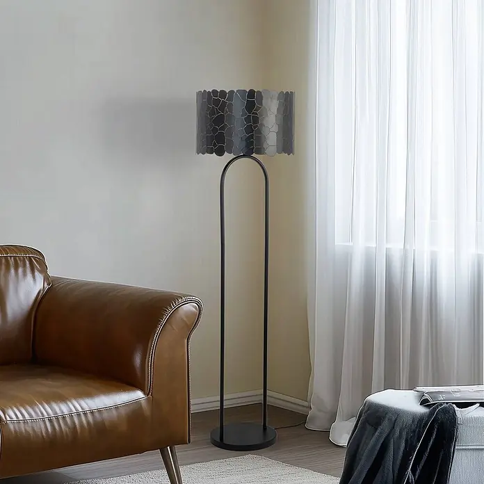 Lindby Stehlampe Wohnzimmer in schwarz, gold, Stehleuchte -  - Schwarze Stehlampe mit dunklem, strukturiertem Lampenschirm und geschwungenem Gestell, neben einem braunen Ledersofa.