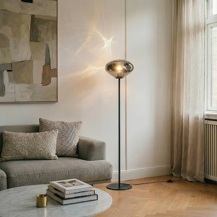 Lindby Stehlampe Wohnzimmer in rauchgrau, schwarz, Stehleuchte -  - Moderne Stehlampe mit Rauchglas-Schirm und schwarzem Stand, die ein Wohnzimmer beleuchtet und dekorative Lichtmuster an die Wand wirft.