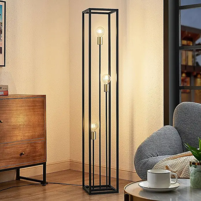 Lindby Stehlampe Wohnzimmer in schwarz matt, Stehleuchte -  - Hohe Stehlampe aus schwarzem Metall mit drei sichtbaren Glühbirnen, beleuchtet eine Raumecke neben Sessel und Schrank.