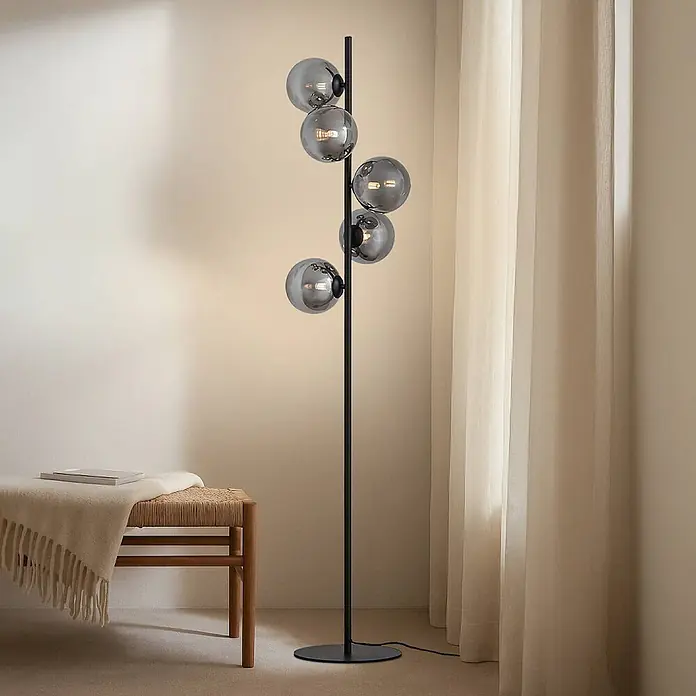 Lindby Stehlampe Wohnzimmer in schwarz, Stehleuchte -  - Eine schwarze Stehlampe mit fünf Rauchglas-Kugel-Schirmen beleuchtet einen gemütlichen Raum neben einer Holzbank und Vorhängen.