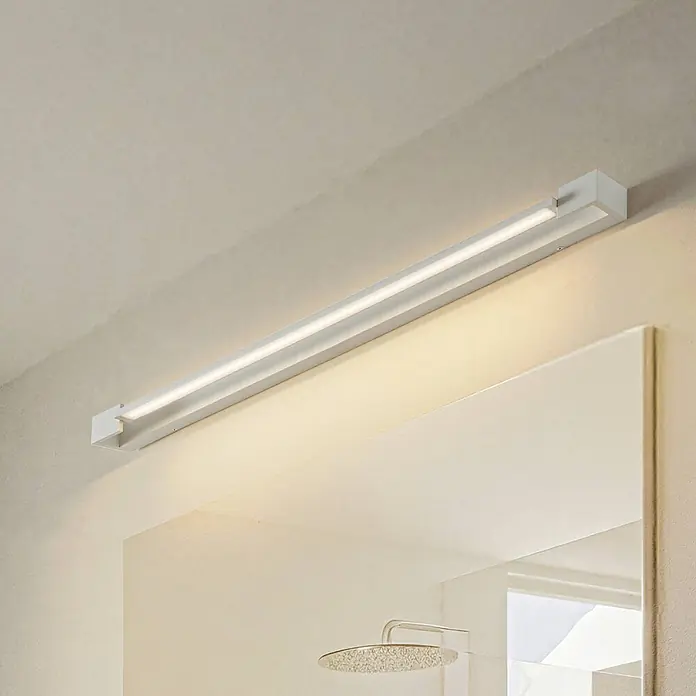 Arcchio LED Wandleuchte innen in weiß, Wandlampe -  - Lange, weiße LED-Spiegelleuchte über einem Spiegel, die den Badbereich warmweiß beleuchtet.