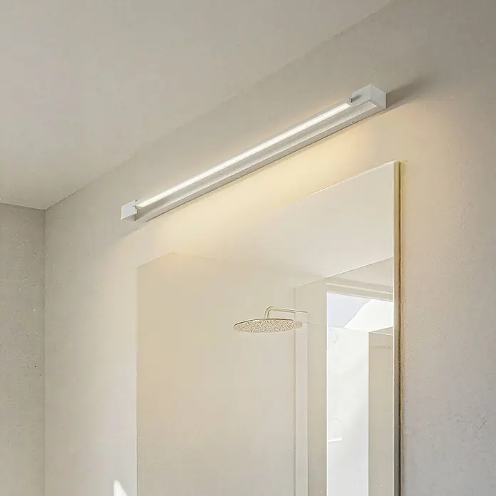 Arcchio LED Wandleuchte innen in weiß, Wandlampe -  - Weiße, lineare LED-Spiegelleuchte über einem Spiegel im Badezimmer installiert. Eine Duschbrause spiegelt sich wider.