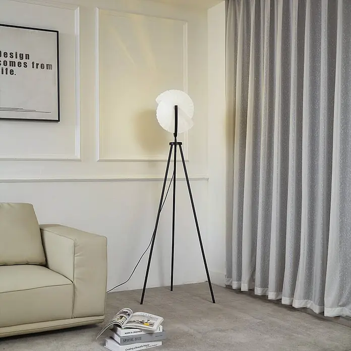 Lucande Stehleuchte Helva - schwarz, weiß / opal, E27 - Eine schwarze Dreibein-Stehlampe mit weißem Faltenlampenschirm steht neben einem beigefarbenen Sofa und spendet Licht.