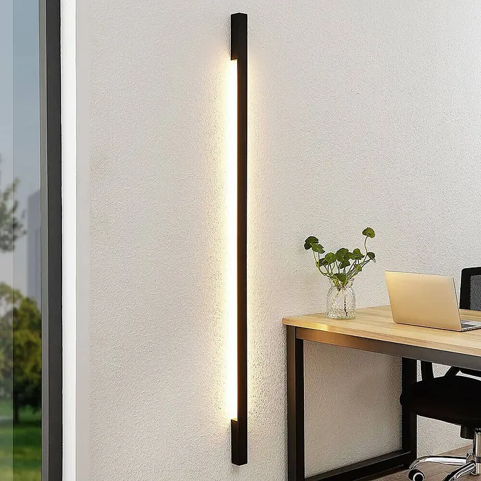 Arcchio LED Wandleuchte innen in schwarz, Wandlampe -  - Eine elegante, schwarze vertikale LED-Wandleuchte beleuchtet eine strukturierte weiße Wand, neben einem Schreibtisch mit Laptop und Pflanze.