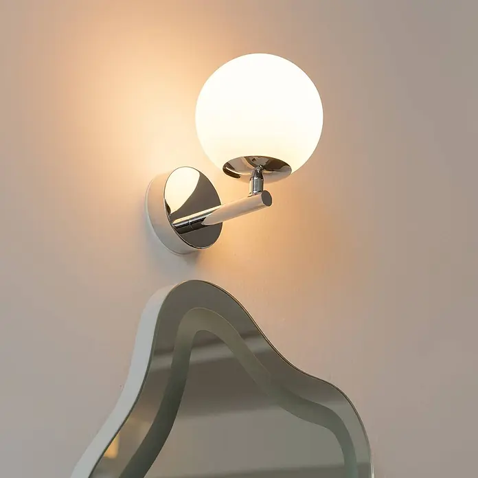 Arcchio LED Wandleuchte innen in chrom, weiß, Wandlampe -  - Eine moderne Wandleuchte mit kugelförmigem weißem Glasschirm und Chromfuß, die warmes Licht auf eine Wand über einem Wellenspiegel wirft.
