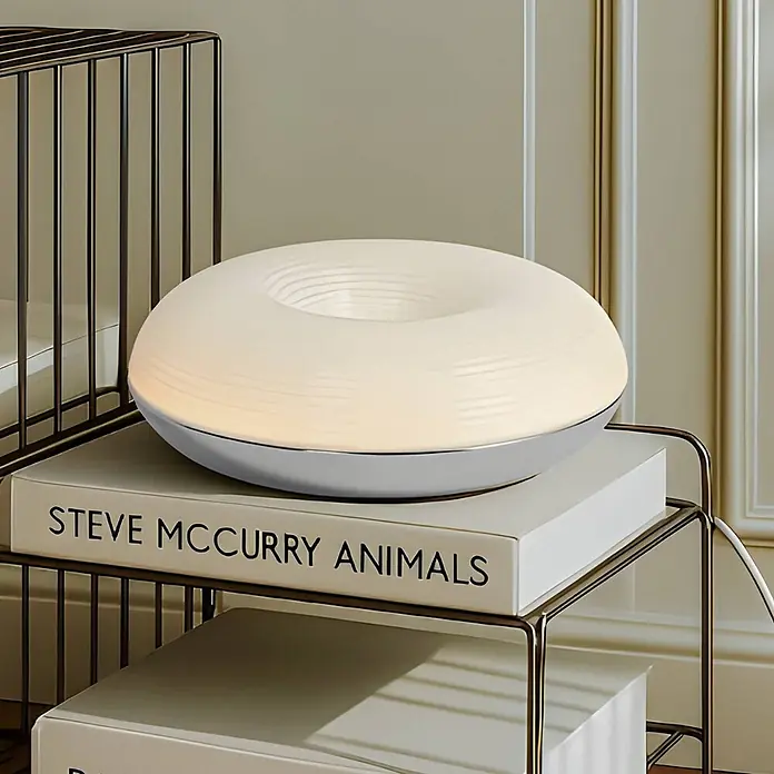 Lindby LED Wandleuchte innen in weiß, chrom, dimmbar Wandlampe -  - Beleuchtete weiße, ringförmige Lampe mit silbernem Sockel, auf einem Buch auf einem Metallregal.
