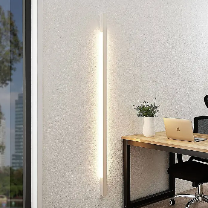 Arcchio LED Wandleuchte innen in weiß, Wandlampe -  - Hohe weiße LED-Wandleuchte, vertikal an einer Wand montiert, beleuchtet einen modernen Schreibtischbereich mit Laptop und Pflanze.