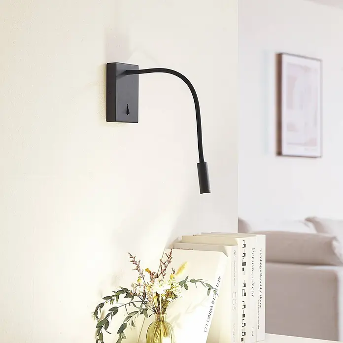Lucande LED Wandleuchte innen in schwarz, Leselampe Wand -  - Eine schwarze Wandleseleuchte mit flexiblem Arm, über Büchern und einer Pflanze in einem Wohnambiente.