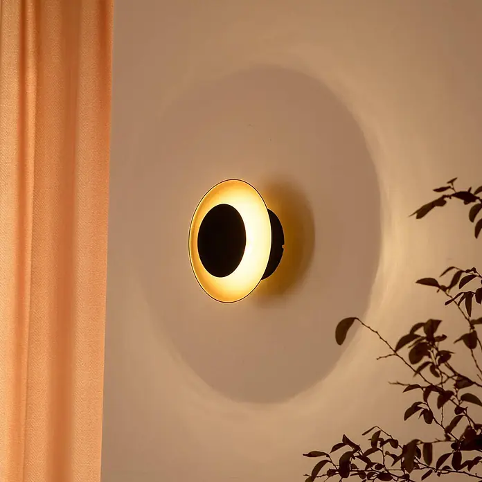 Lindby Wandleuchte Tiama - schwarz - Moderne schwarze und goldene runde Wandleuchte wirft warmes Licht auf eine Wand, daneben ein orangefarbener Vorhang.