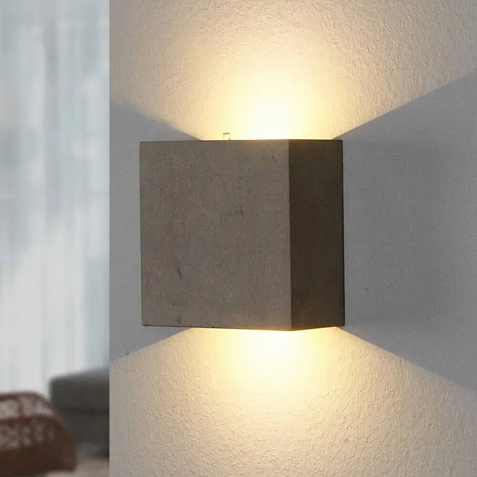 Lindby Wandleuchte Yva - alu / grau / zink - Graue kubische Wandlampe an einer strukturierten Wand, die warmes Licht nach oben und unten wirft.