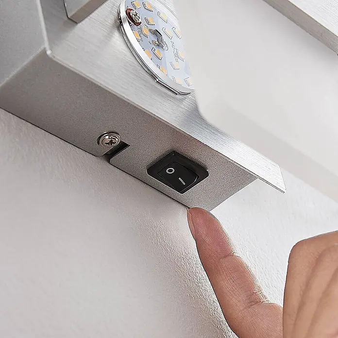 Lindby Wandleuchte Yorick - alu / grau / zink, weiß / opal - Finger drückt schwarzen Wippschalter an silberner gebürsteter Metall-LED-Leuchte.