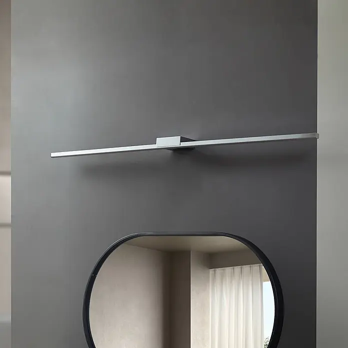 Lucande LED Wandleuchte innen in chrom, Wandlampe -  - Moderne lineare silberne Leuchte an einer grauen Wand über einem ovalen Spiegel montiert.
