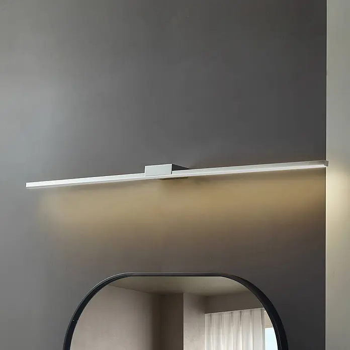 Lucande LED Wandleuchte innen in chrom, Wandlampe -  - Schlanke lineare weiße LED-Spiegellampe über einem Spiegel, wirft warmes Licht auf eine graue Wand.