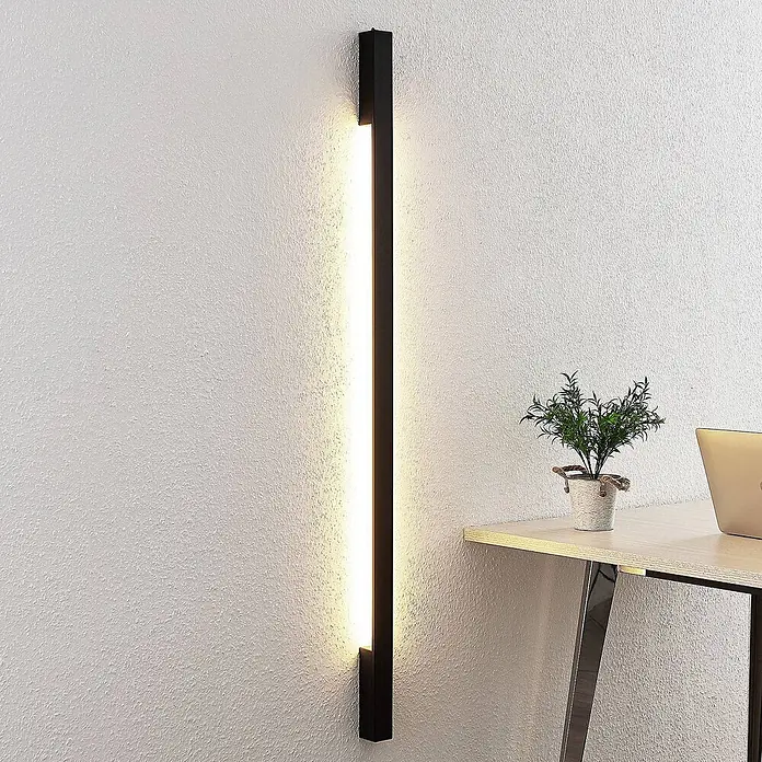Arcchio LED Wandleuchte innen in schwarz, Wandlampe -  - Eine schwarze, vertikale LED-Wandleuchte ist an einer weißen Wand montiert und wirft warmes Licht neben einen Schreibtisch mit Pflanze und Laptop.