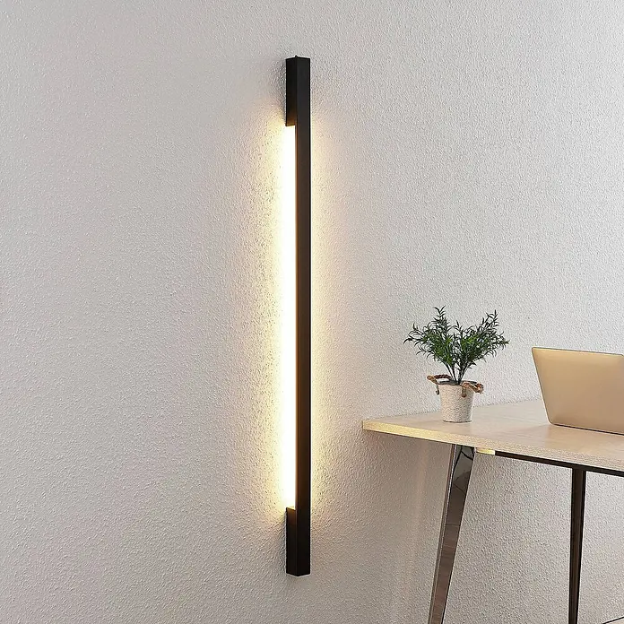 Arcchio LED Wandleuchte innen in schwarz, Wandlampe -  - Lange, schmale, schwarze LED-Wandleuchte vertikal an einer strukturierten Wand, die den Bereich über einem hölzernen Schreibtisch mit einer Pflanze beleuchtet.