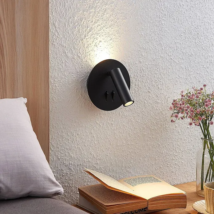 Lucande LED Wandleuchte innen in schwarz matt, Wandlampe -  - Schlanke schwarze Wandleseleuchte mit zwei Schaltern, beleuchtet ein offenes Buch auf einem Nachttisch vor einer strukturierten weißen Wand.
