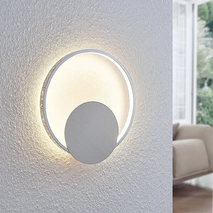 Lindby Wandlampe, Wandleuchte innen, Innenleuchte Wand, inkl. 11,2W LED -  - Eine moderne, silberne runde LED-Wandleuchte mit versetzter Innenscheibe, an einer strukturierten Wand montiert.