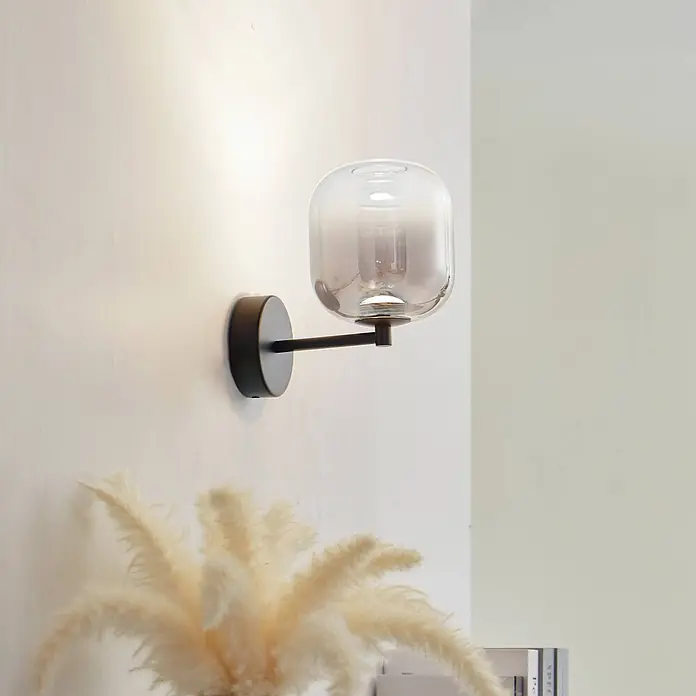 Lucande Wandleuchte Avelina - schwarz, alu / grau / zink - Schwarze Wandleuchte mit Rauchglas-Schirm, beleuchtet eine Wand. Pampasgras dekoriert den Vordergrund.