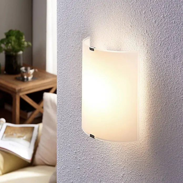 Lindby LED Wandleuchte innen in transparent satiniert, Wandlampe -  - Eine gebogene Wandleuchte aus satiniertem Glas ist an einer strukturierten Wand montiert und spendet warmes Licht im Wohnbereich.
