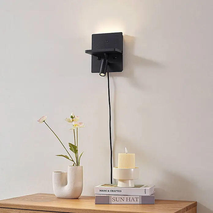 Lindby Wandleuchte Auden - schwarz, schwarz - Schwarze Wandlampe mit integriertem Regal und verstellbarem Spotlicht, eingesteckt und eine Wand beleuchtend. Über einer Holzfläche montiert.