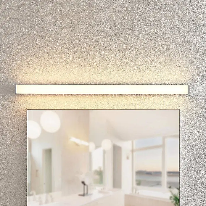 Lindby LED Wandleuchte innen in weiß, chrom, Wandlampe -  - Eine lange, schmale LED-Spiegelleuchte mit silbernem Rahmen beleuchtet eine strukturierte Wand über einem Badezimmerspiegel.
