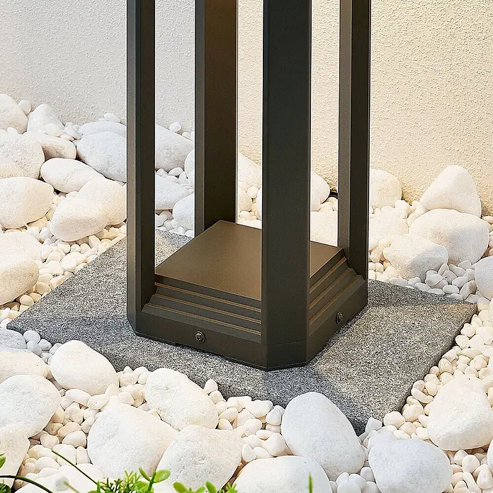 Lucande LED Wegeleuchte Sockelleuchte 50 cm, Gartenlampe, Außenlampe -  - Dunkelgrauer Sockel einer Außenleuchte auf einer grauen Platte, umgeben von weißen Kieselsteinen für Gartenbeleuchtung.