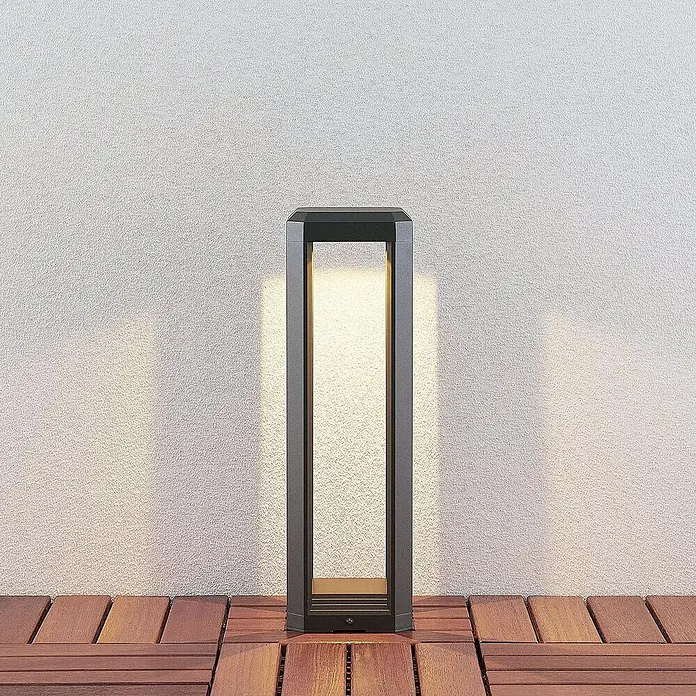 Lucande LED Wegeleuchte Sockelleuchte 50 cm, Gartenlampe, Außenlampe -  - Moderne dunkelgraue Außenpollerleuchte auf Holzterrasse, die warmes Licht auf eine strukturierte Wand wirft.