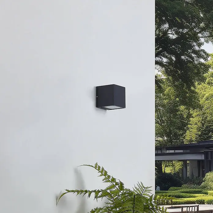 Lucande LED Wandleuchte aussen in dunkelgrau, up down Aussenlampe Wand -  - Schwarze, würfelförmige Außenwandleuchte an einer Außenwand. Mit Grün und einem Gebäude im Hintergrund.