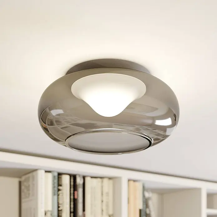 Lucande Deckenleuchte Mijo - alu / grau / zink - Moderne Deckenleuchte mit Rauchglasschirm und weißem, konischem Diffusor, beleuchtet einen Raum mit Bücherregalen.