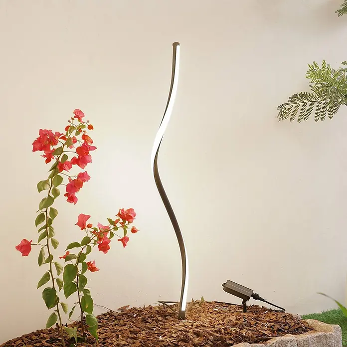 Lindby Erdspießleuchte Sabiel -  - Moderne S-förmige dunkle LED-Gartenwegleuchte beleuchtet rote Blumen in einem Gartenbeet mit Holzspänen.
