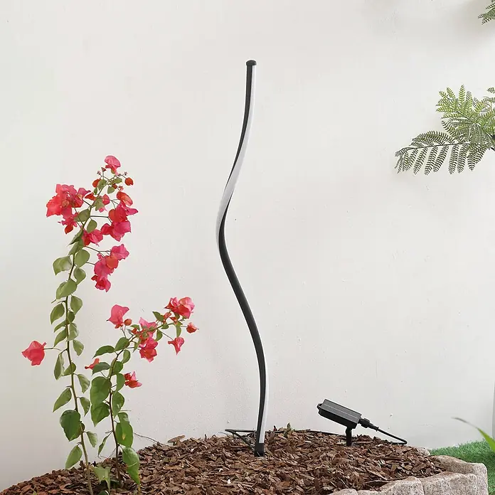 Lindby Erdspießleuchte Sabiel -  - Eine schwarze, gebogene LED-Gartenleuchte mit Solarpanel in einem Blumenbeet mit roten Blüten.
