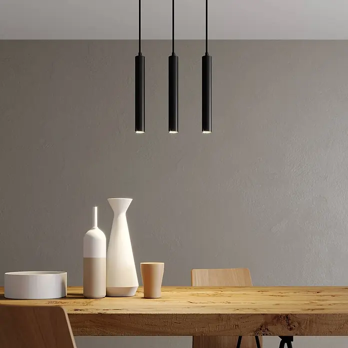 Arcchio LED Pendelleuchte in schwarz, Hängelampe -  - Drei schwarze zylindrische Pendelleuchten hängen über einem Holztisch und beleuchten Dekoartikel.