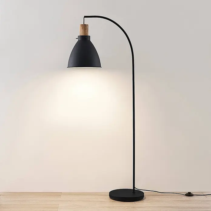 Lindby Stehlampe Skandi Holz Metall, Leselampe Stehlampe Wohnzimmer -  - Eine schwarze Stehlampe mit Holzdetail am Schirm beleuchtet eine helle Wand und steht auf Holzfußboden.