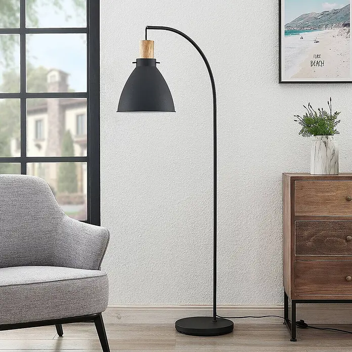 Lindby Stehlampe Skandi Holz Metall, Leselampe Stehlampe Wohnzimmer -  - Schwarze Bogen-Stehleuchte mit Holzdetail am Schirm, im Wohnzimmer neben einem grauen Sessel platziert.
