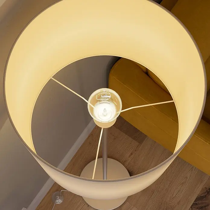 Lindby Stehlampe Wohnzimmer in weiß, nickel matt, Stehleuchte -  - Beleuchtete Stehlampe mit beigem Stoffschirm und sichtbarer Glühbirne, beleuchtet einen Bereich mit gelbem Sofa und Holzboden.
