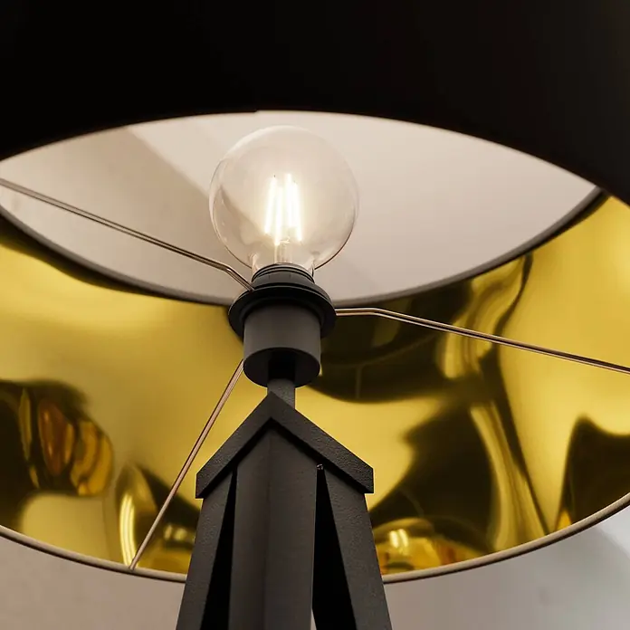 Lindby Stehlampe Dreibein schwarz gold mit Fußschalter, ohne Leuchtmittel -  - Nahaufnahme einer Lampe mit schwarzem Schirm, goldenem Inneren und leuchtender klarer LED-Birne.