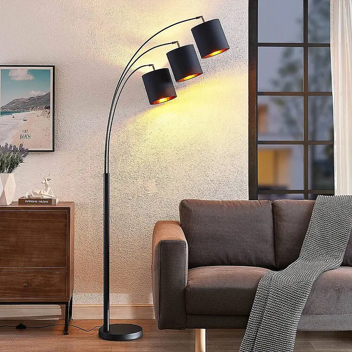 Lindby Stehlampe Wohnzimmer in schwarz, gold, Stehleuchte -  - Schwarze Bogenlampe mit drei Armen und zylindrischen Schirmen, spendet warmes Licht neben einem Sofa.