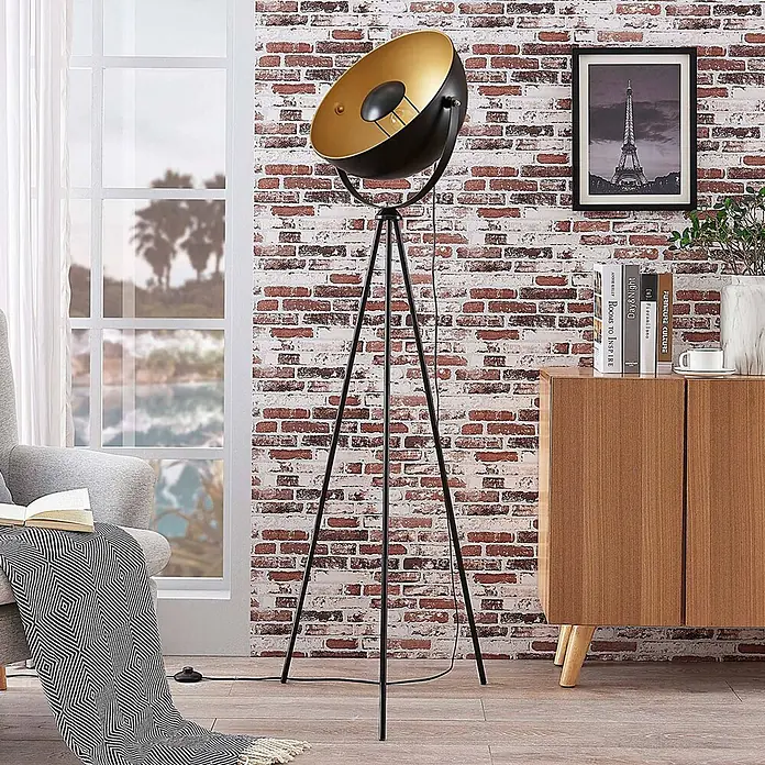 Lindby Tripod / Dreibein Stehlampe Schwarz Gold, ohne Leuchtmittel -  - Eine moderne schwarze Stativ-Stehlampe mit goldfarbenem Reflektor steht in einem Wohnzimmer auf einem Holzboden.
