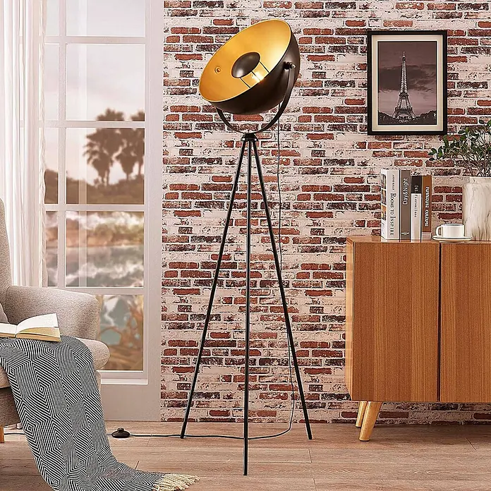 Lindby Tripod / Dreibein Stehlampe Schwarz Gold, ohne Leuchtmittel -  - Große schwarze und goldene Stativ-Stehlampe beleuchtet ein modernes Wohnzimmer mit Ziegelwand.