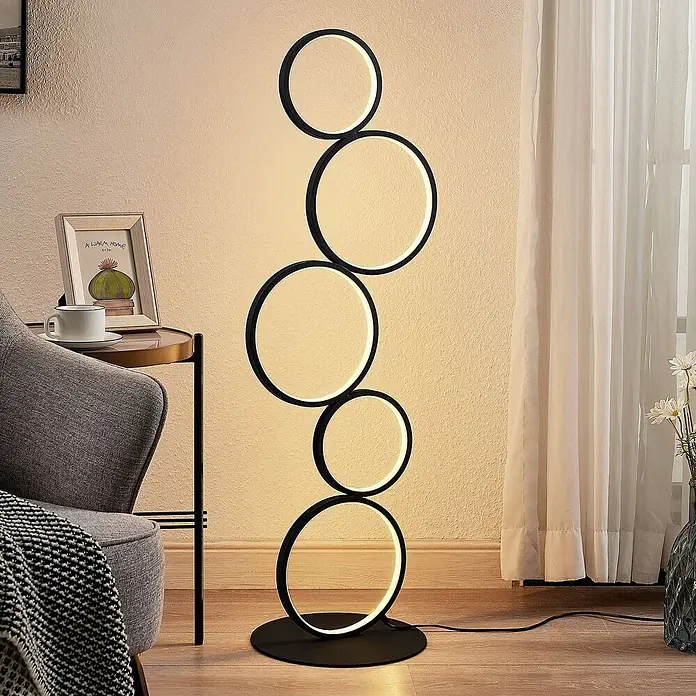 Lindby LED Stehlampe Wohnzimmer in schwarz, Stehleuchte -  - Moderne schwarze Stehlampe mit fünf beleuchteten LED-Ringen, auf Holzboden in gemütlichem Wohnzimmer neben Sessel.