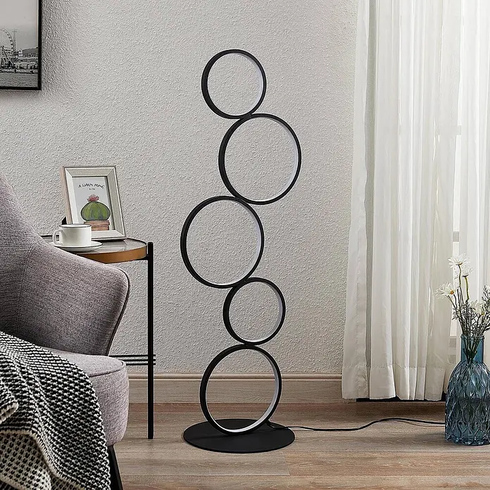 Lindby LED Stehlampe Wohnzimmer in schwarz, Stehleuchte -  - Moderne schwarze Stehleuchte mit fünf beleuchteten LED-Ringen, aufrecht stehend im Wohnzimmer neben einem grauen Sessel.