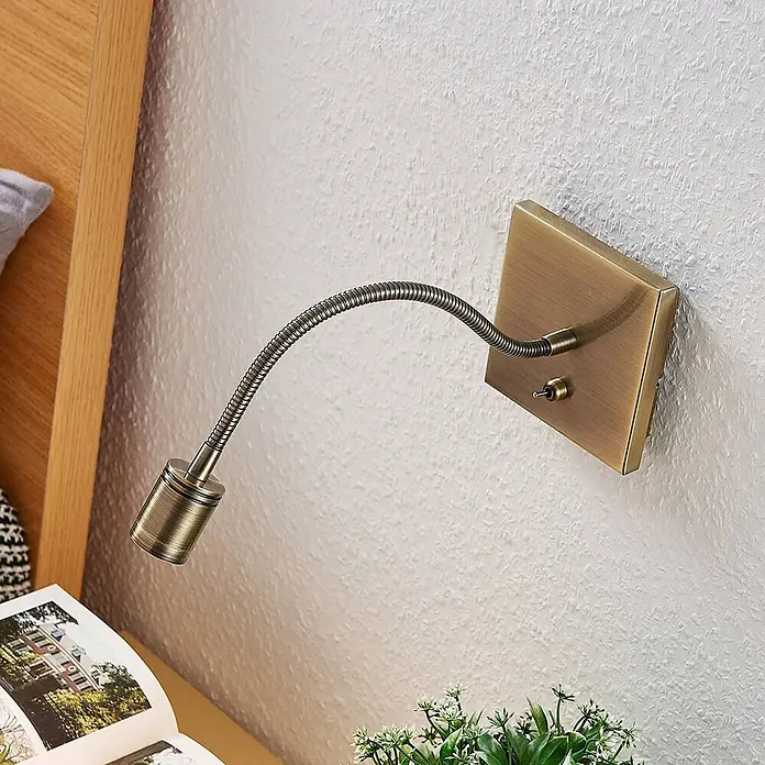 Lindby LED Wandlampe biegsamer Flexarm, Strahler Schlafzimmer, Küche -  - Messingfarbene Wand-Leselampe mit flexiblem Schwanenhals, die ein Buch neben einem hölzernen Bettrahmen beleuchtet.
