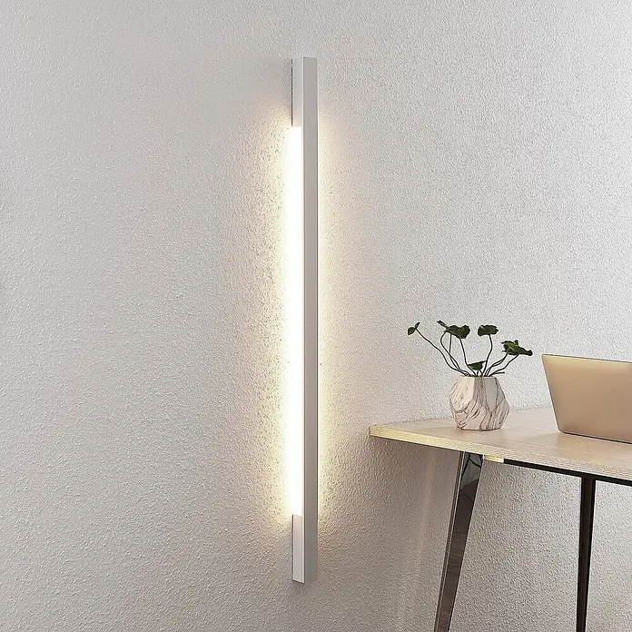 Arcchio LED Wandleuchte innen in weiß, Wandlampe -  - Eine schlanke, weiße LED-Wandleuchte vertikal montiert, spendet Umgebungslicht neben einem Holzschreibtisch mit Pflanze und Laptop.