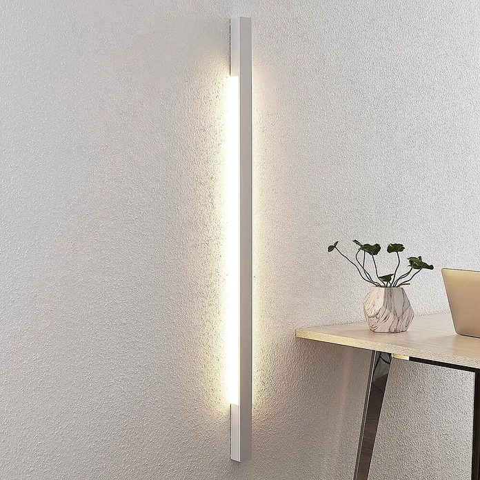 Arcchio LED Wandleuchte innen in weiß, Wandlampe -  - Weißes vertikales LED-Wandlicht, an einer strukturierten Wand montiert, beleuchtet den Bereich neben einem Holzschreibtisch.
